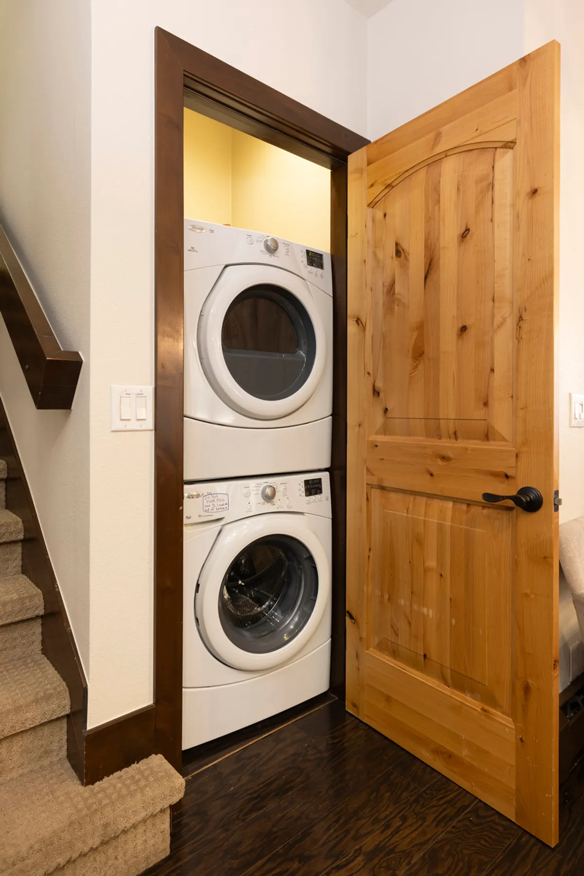 The photo highlights a laundry area adjacent to a wooden door and staircase, with a washer, dryer, and visible storage space. The setting feels practical and homey, reinforcing the convenience of organizing gear and essentials after outdoor activities.