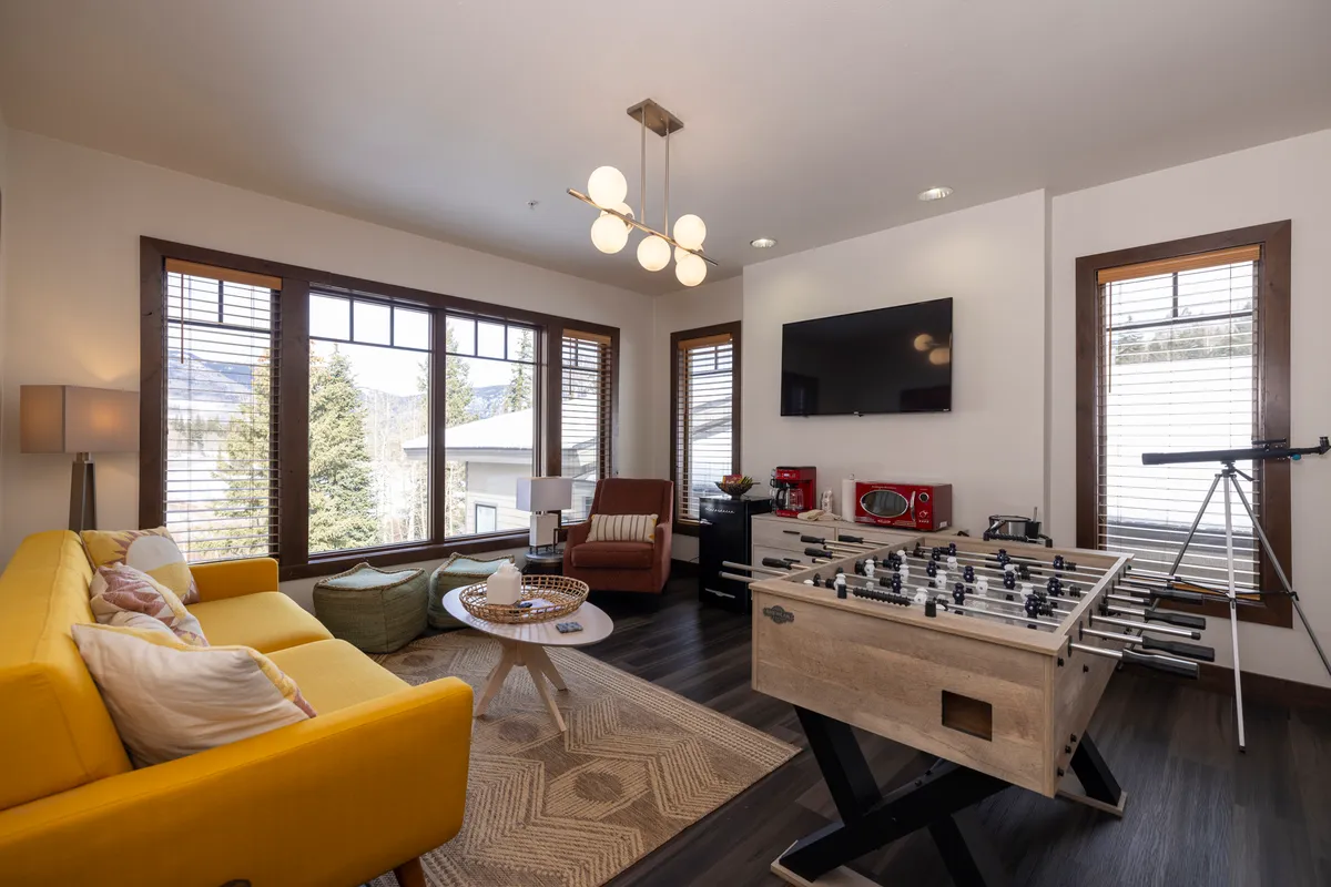 A cheerful interior game area featuring a foosball table, vibrant yellow sofa, and large windows inviting in natural light. The space is inviting for both friendly competition and casual relaxation, perfect for family game nights or lively gatherings.