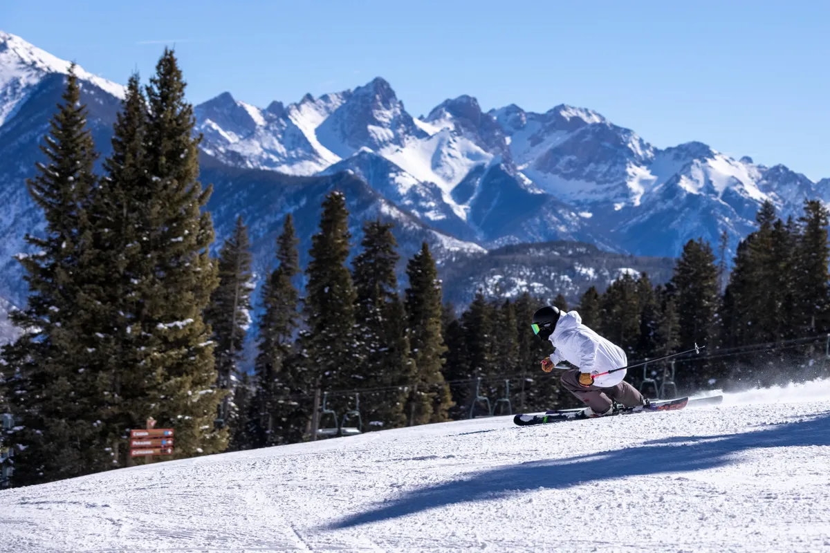 A skier glides down a pristine, snow-covered slope framed by pine trees, with the majestic mountain range stretching across the background beneath a clear sky. The scene captures the adventurous spirit and breathtaking natural beauty of the San Juan Mountains, inviting viewers to imagine the outdoor activities available just moments from the property.