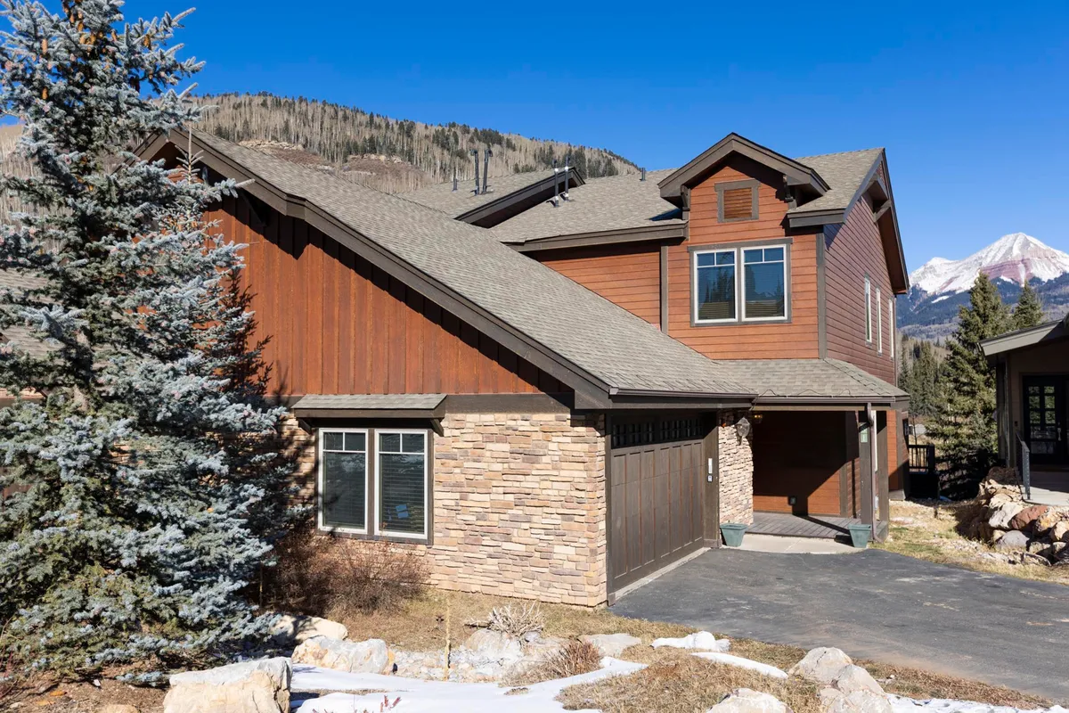 This exterior shot displays the townhome's wooden siding, stone accents, and large windows, set against a picturesque mountain backdrop. The inviting façade hints at both comfort and adventure, while the spacious outdoor area visually connects to activities like outdoor dining and family gatherings.