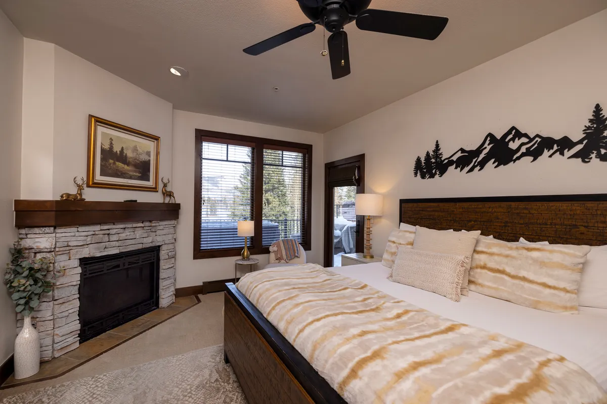 A serene bedroom featuring a cozy bed next to a fireplace, a large window letting in soft natural light, and tasteful mountain-inspired decor. The ambiance is peaceful, inviting relaxation after a long day outdoors.
