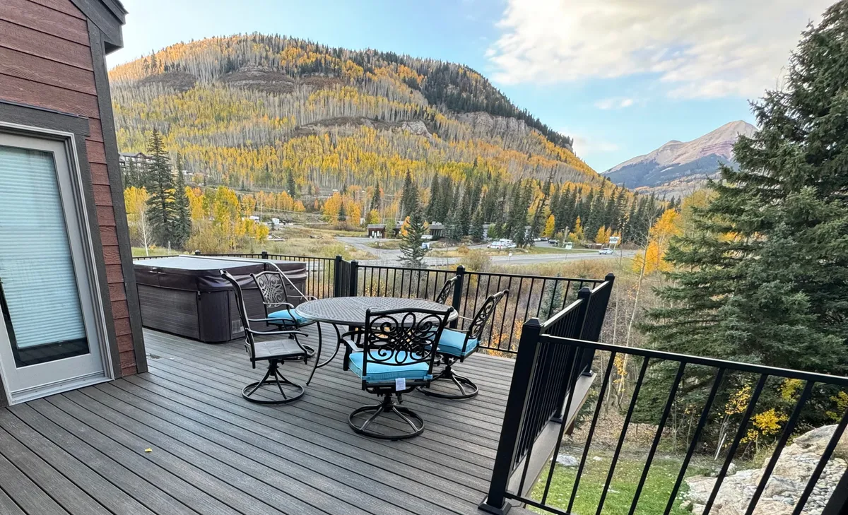 An inviting outdoor seating area with a private hot tub overlooks the mountains, framed by golden autumn foliage. The tranquil setting offers a perfect spot for unwinding after a day of adventure, with panoramic views and a sense of closeness to nature.