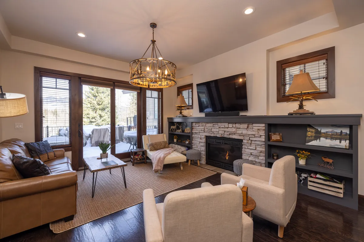 A cozy living room featuring a stone fireplace, plush sofa and armchairs, a large chandelier, and expansive windows that frame the mountain scenery. Sunlight streams in, illuminating the warm, inviting space and drawing attention to the relaxing and scenic environment found within the home.