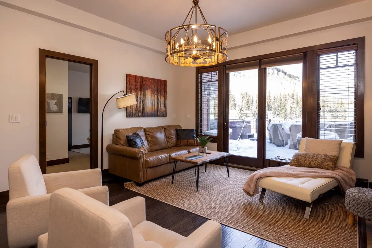 A comfortable living room with a leather sofa, large windows providing mountain views, and warm ambient lighting from a chandelier. The inviting atmosphere is perfect for peaceful reflection and enjoying the surrounding alpine scenery from inside.