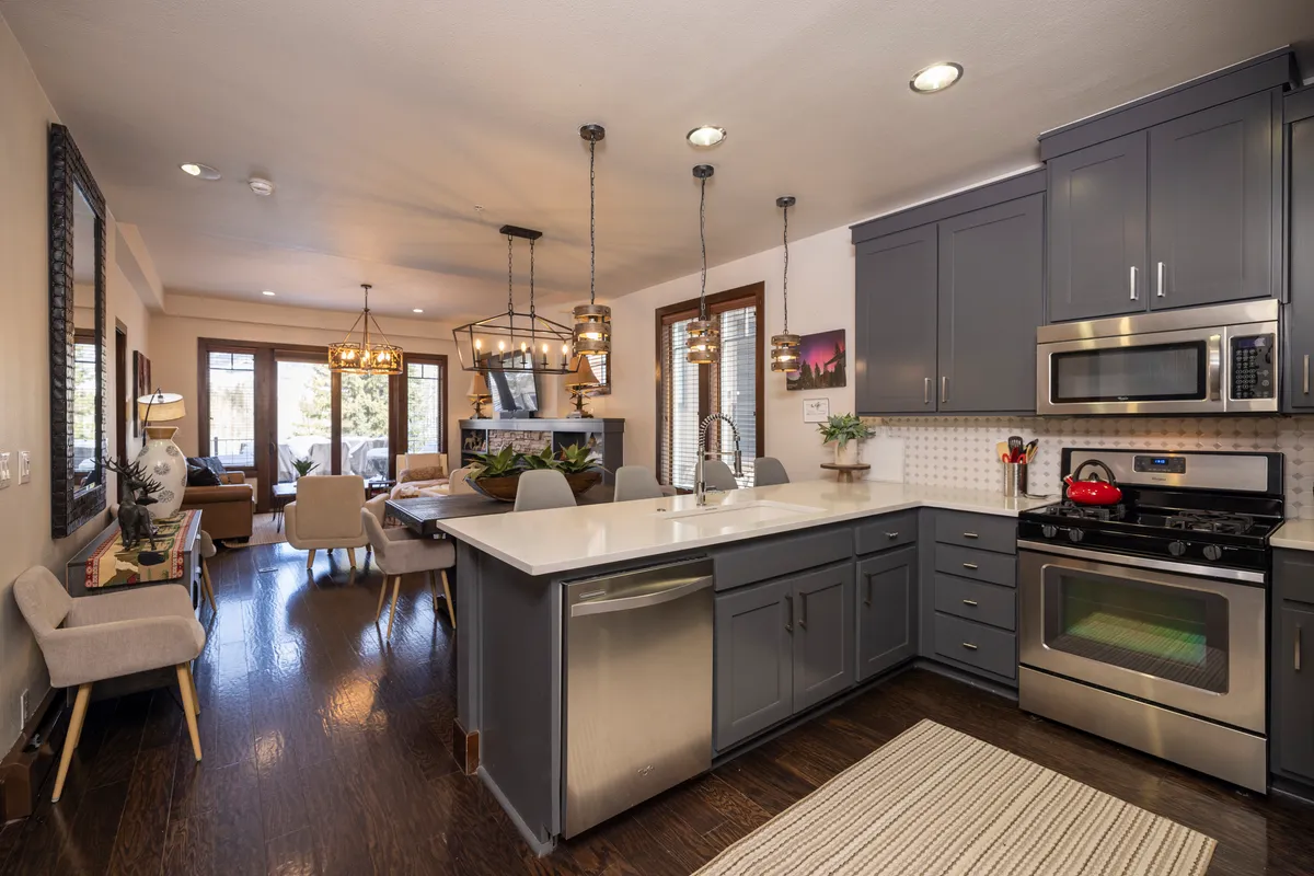 The image showcases a spacious, modern kitchen with a large island, gleaming stainless steel appliances, a cozy dining area, and expansive windows flooding the space with natural light. The inviting setup hints at gatherings, celebrations, and relaxed meals with mountain scenery just beyond the glass.