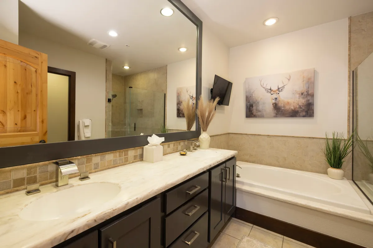 A spacious bathroom with a double vanity featuring a marble countertop, a framed mirror, a bathtub, decorative wall art, and a separate shower area. The colors and decor evoke a tranquil, spa-like feel.