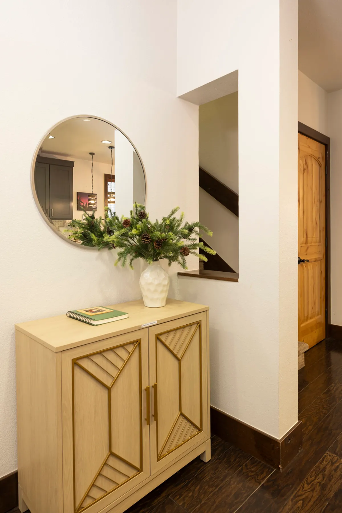 This image captures an inviting entryway featuring a decorative cabinet, round mirror, potted greenery, and a staircase. The space is clean, organized, and welcoming, suggestive of a well-designed transition area between the outdoors and the main living spaces.