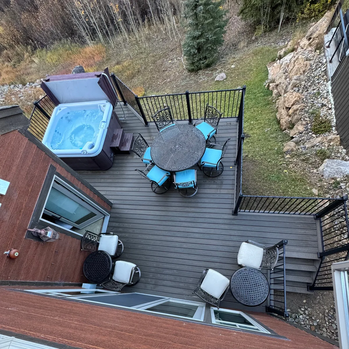 An outdoor deck featuring a round dining table and a hot tub, surrounded by greenery and bordered by a deck railing. The setting invites al fresco dining, relaxation, and social gatherings with views of the natural landscape.