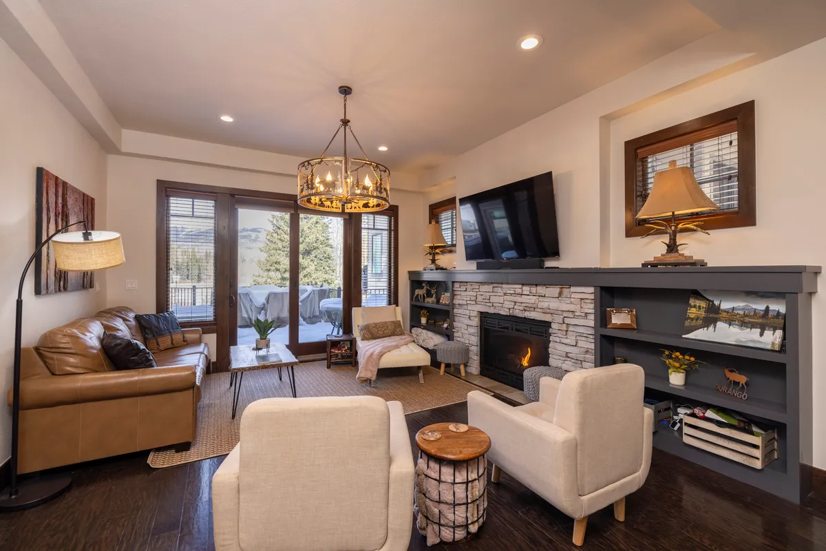 A warm living room featuring a stone fireplace, cozy sofa, large windows that frame serene mountain views, and a coffee table positioned for conversation or games. The natural light enhances the inviting atmosphere, making it an ideal spot for relaxation and socializing after a day outdoors.