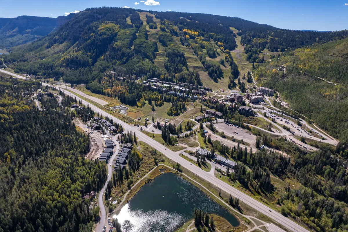 A sweeping view captures mountain slopes, a sparkling lake, residential buildings, and a network of roadways. The foreground features Twilight Lake, while the background is dominated by forested mountains, suggesting year-round outdoor activity. The scene hints at both summer water sports and winter snow adventures, with the community nestled at the lake's edge.
